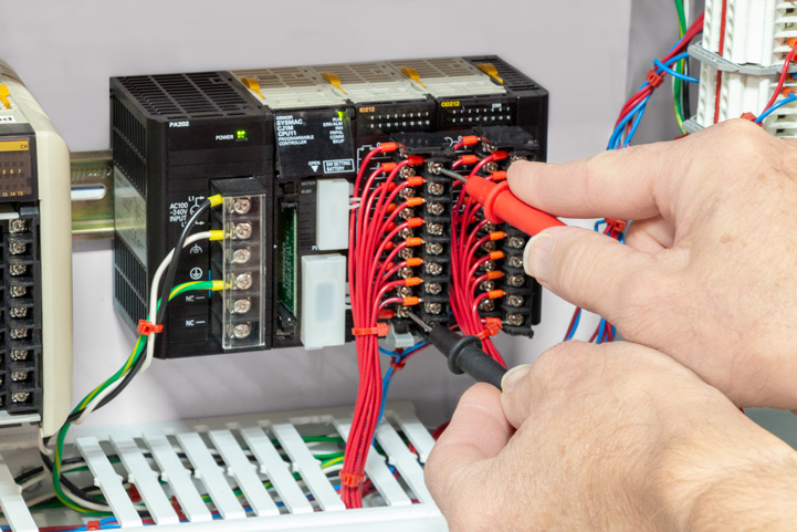Technician using multimeter probes to test connections on an Omron SYSMAC CJ-series PLC module in an industrial control cabinet. The image shows organized red wiring connected to terminal blocks on the CJ-series input/output modules
