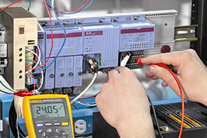 B&R System 2003 testing systems Classic Automation Technician measuring electrical components with a multimeter, working on a panel with multiple controllers and wires.